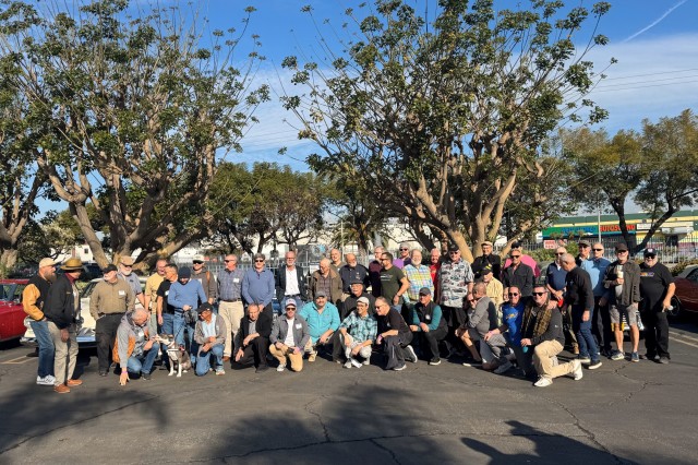Forty-two men in a group photo, some standing, some kneeling, in a parking lot with trees in the background and classic cars on either side of them.